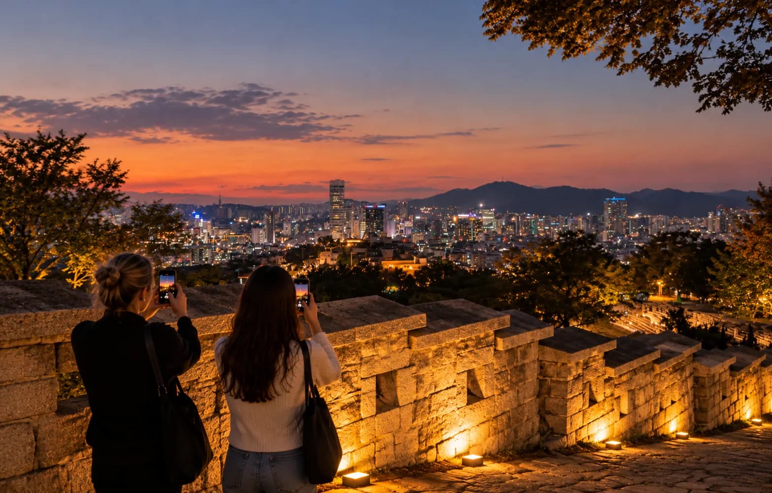 Two people photographing the city skyline at dusk from the Naksan section of the Seoul City Wall
